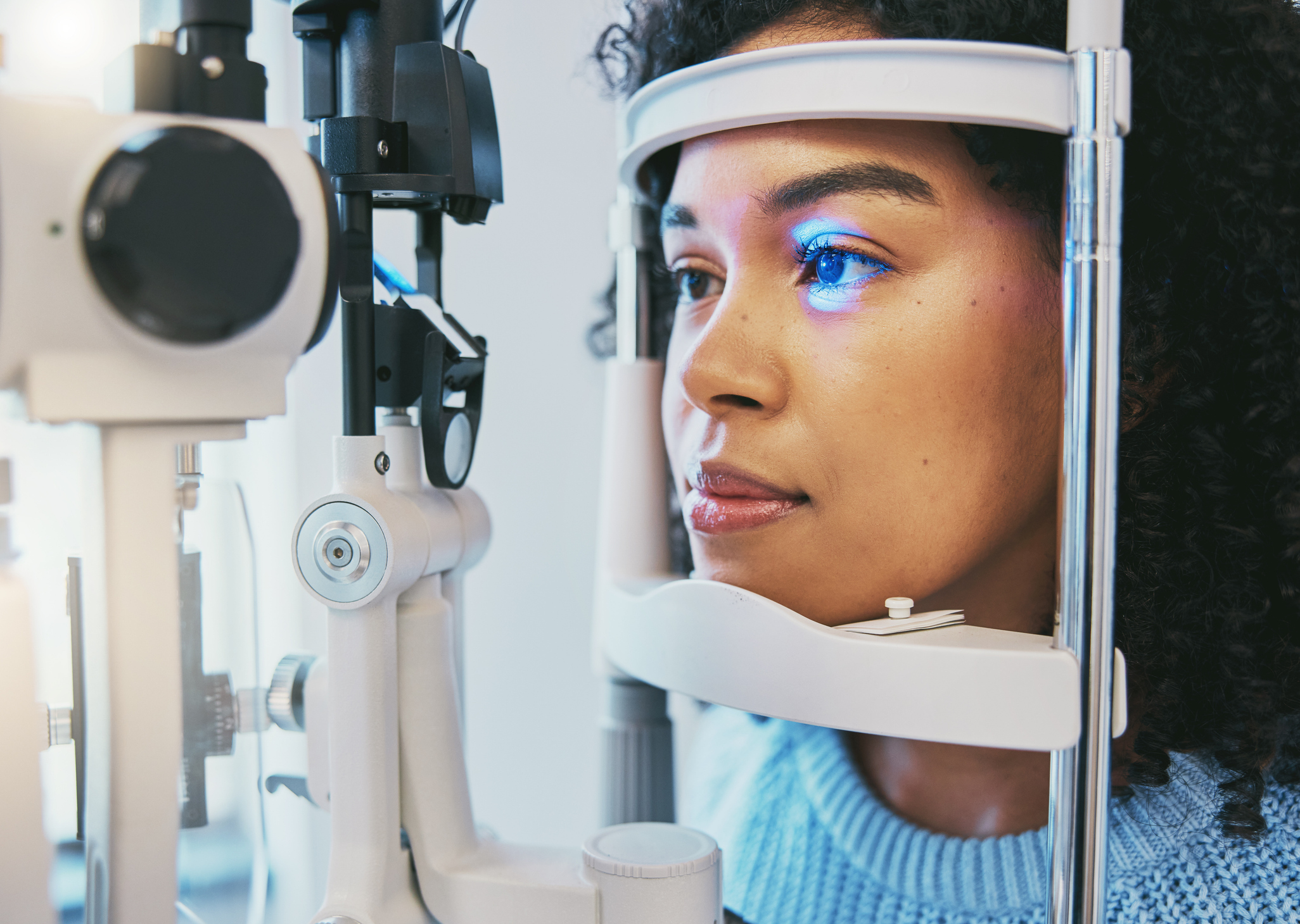 A woman places her face on the headrest of a slit lamp for an eye exam.