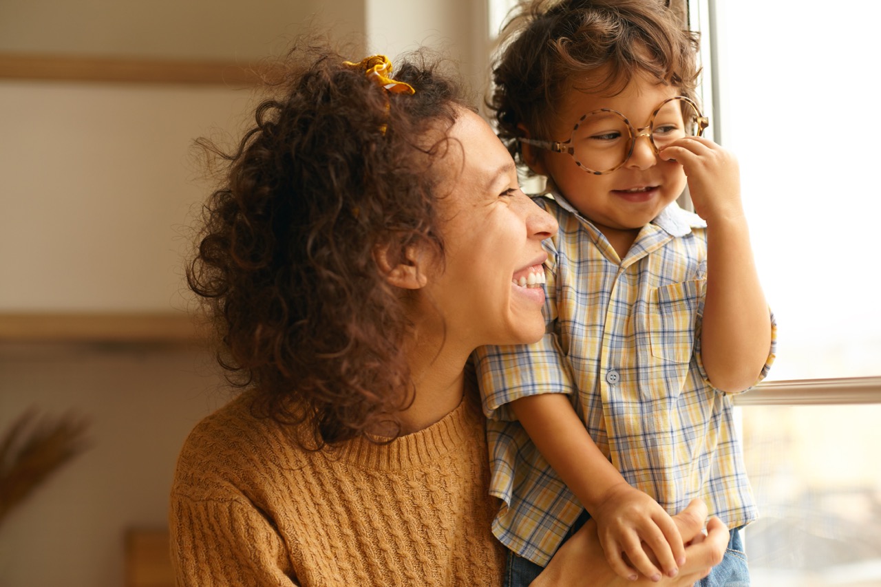 A smiling mother hugs her toddler, who is wearing glasses.