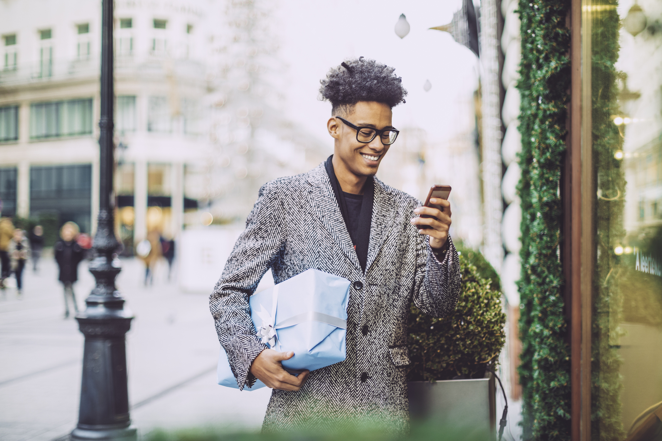 A man wearing glasses stands outside in cold weather. He is wearing a coat and carrying a wrapped package.